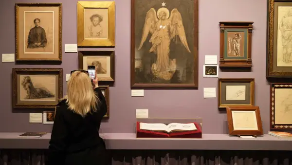 Visite d'un salon du dessin : une femme photographie une œuvre d'art, entourée de nombreux croquis et peintures exposés sur un mur lilas.