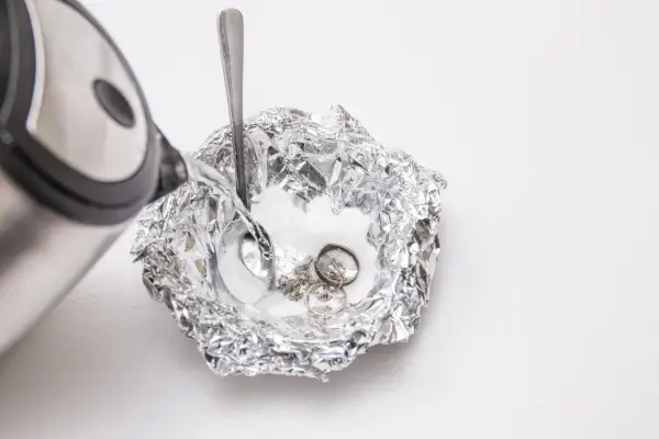 Nettoyage d'argenterie noircie : de l'eau chaude est versée sur des bijoux en argent et une cuillère dans une coupelle en papier aluminium.