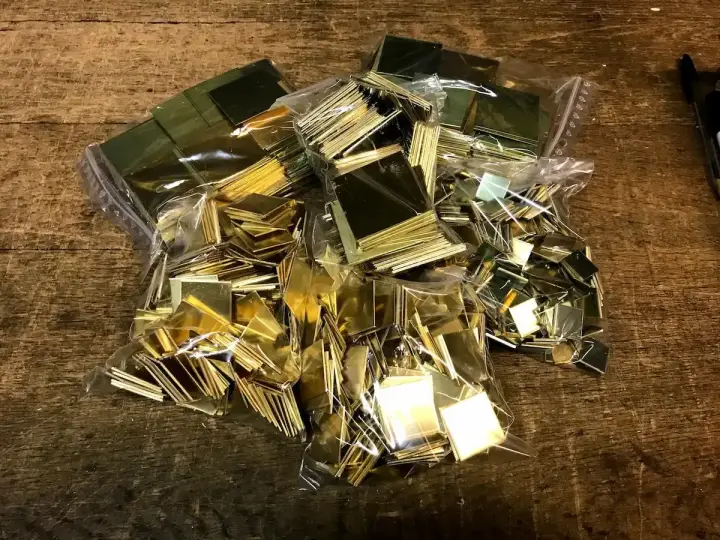 Des petits carrés de laiton brillants, prêts pour un projet de comment décaper du laiton. Ils sont dans des sacs en plastique sur une table en bois.