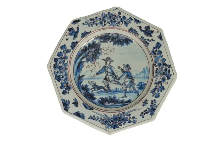 Assiette octogonale en céramique de Marseille, décorée de deux hommes et d'un chien dans un paysage champêtre, bordée de motifs floraux bleus.