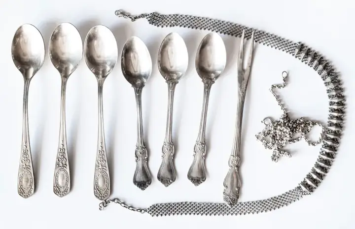 Six cuillères et une fourchette anciennes, ornées, sont disposées avec une chaîne argentée. Idéal pour apprendre comment nettoyer l'argenterie.
