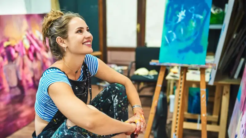 Une artiste rêveuse, les mains tachées de peinture, s'interroge sur comment devenir artisan d'art. Son atelier est rempli de toiles colorées.