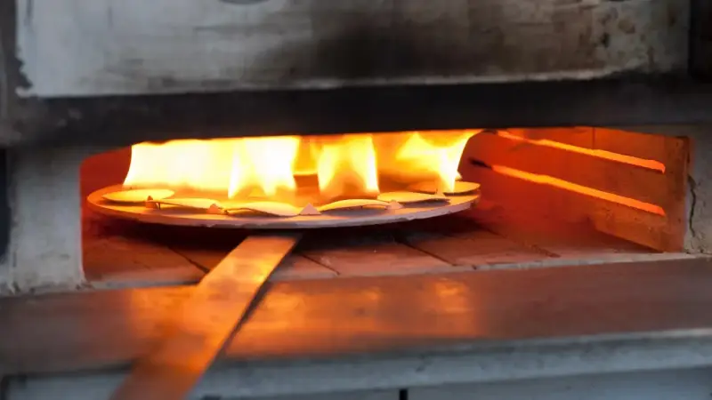 Des pièces de métal attendent d'être émaillées dans un four, sous des flammes vives. L'artisan émailleur surveille attentivement la cuisson.